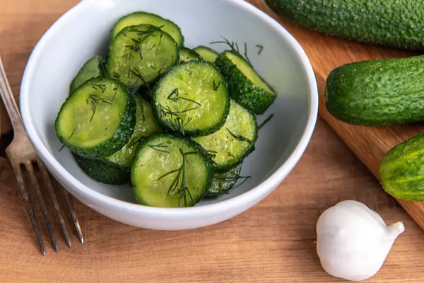 Ahşap bir masada seramik beyaz bir kasede organik salatalıklar dilimler. Taze sebze ve dereotu, ahşap dokuma masasında sarımsak. Salata..