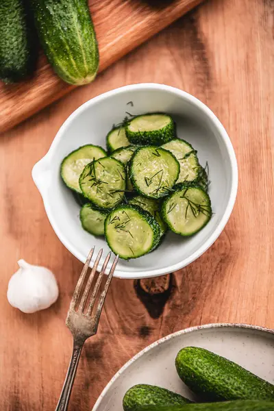 Ahşap bir masada seramik beyaz bir kasede organik salatalıklar dilimler. Taze sebze ve dereotu, ahşap dokuma masasında sarımsak. Salata..