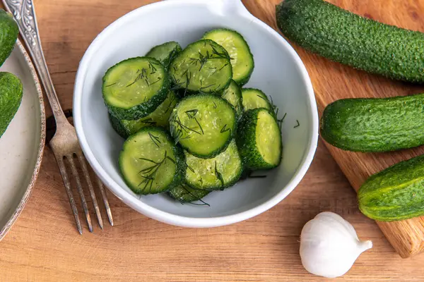 Ahşap bir masada seramik beyaz bir kasede organik salatalıklar dilimler. Taze sebze ve dereotu, ahşap dokuma masasında sarımsak. Salata..