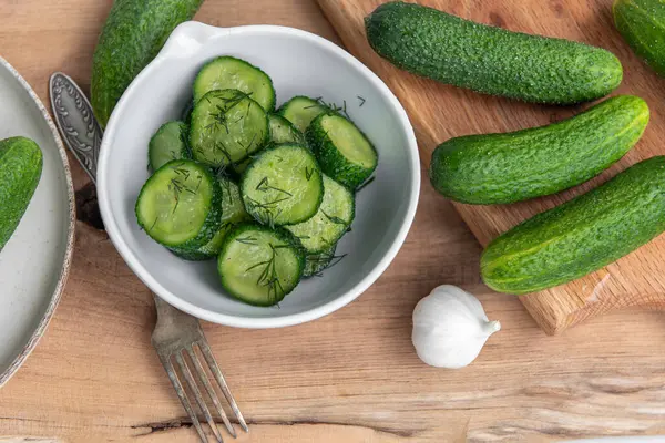 Ahşap bir masada seramik beyaz bir kasede organik salatalıklar dilimler. Taze sebze ve dereotu, ahşap dokuma masasında sarımsak. Salata..