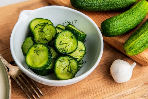 Ahşap bir masada seramik beyaz bir kasede organik salatalıklar dilimler. Taze sebze ve dereotu, ahşap dokuma masasında sarımsak. Salata..