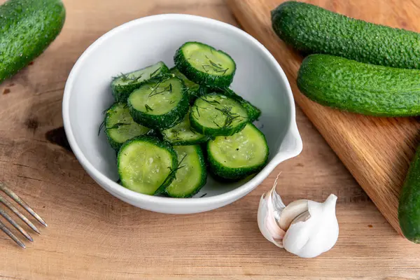 Ahşap bir masada seramik beyaz bir kasede organik salatalıklar dilimler. Taze sebze ve dereotu, ahşap dokuma masasında sarımsak. Salata..