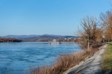 Bir feribot Tuna Nehri 'ni açık bir yay gökyüzünün altında, uzak tepeler ve çıplak ağaçlarla kaplı nehir kenarındaki bir yol ile geçer..