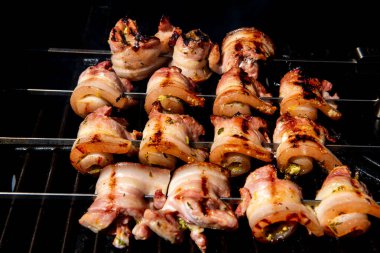 Sulu domuz eti dilimleri sarılmış ve şiş vaziyette açık havada ızgara pişiriliyor. Yaz barbeküsü, açık hava yemekleri ve geleneksel ev yapımı yemek kavramları için mükemmel..