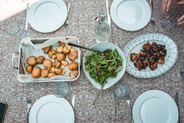 Bir kase taze yeşil salata, kızarmış patates ve ızgara domuz eti şişleri de dahil olmak üzere ev yapımı yemeklerle dolu bir yemek masasının en üst görüntüsü. Kaba bir aile tarzı yemek, masa örtüsünde servis edilir..