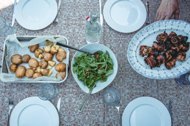 Bir kase taze yeşil salata, kızarmış patates ve ızgara domuz eti şişleri de dahil olmak üzere ev yapımı yemeklerle dolu bir yemek masasının en üst görüntüsü. Kaba bir aile tarzı yemek, masa örtüsünde servis edilir..