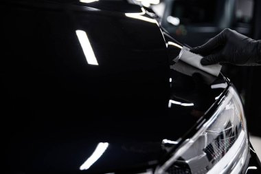 Tamirci siyah eldiven giyerek araba kaputuna seramik kaplama uyguluyor. Detaylı bir stüdyoda, parlak, koruyucu bir kaplama sağlıyor. Ön yüzey araçları üzerinde çalışan ellerin yakın görüntüsü.