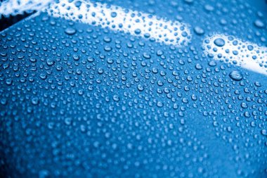 Su damlacıklarının yakın çekimi seramik kaplama uyguladıktan sonra araba yüzeyi üzerindeki hidrofobik etkisini gösteriyor. Görüntü, detaylı bir stüdyoda parlak, iyi korunan mavi bir araba yüzeyi gösteriyor.