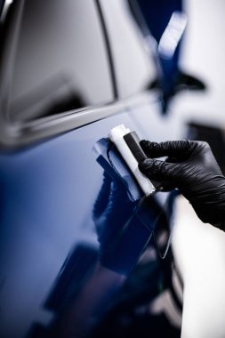 Görüntü, mavi bir arabanın yüzeyine seramik kaplama uygulayan birini gösteriyor. Kişi siyah eldiven giyiyor ve ayarı iyi aydınlatılmış bir atölye ya da garaj gibi görünüyor.