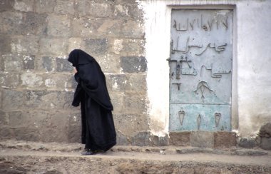 Sanaa 'da geleneksel peçeli bir kadın yürüyor. Geleneksel peçeli Yemenli kadın, Sanaa 'da duvarın yanında süslü süslemeleri olan bir kapıyla yürüyor. 