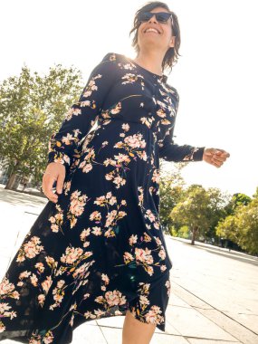 Yazlık elbise ve klasik güneş gözlüğü takan gülümseyen beyaz kadın, Ağaç çizgili bir gezinti güvertesinde yürüyor..