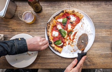 Erkek eller geleneksel Pizzeria 'nın ahşap bir masasında taze pişmiş Napoli Margherita pizzası kesiyor..