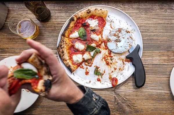 Erkek el, geleneksel Pizzeria 'daki ahşap bir masada taze pişmiş Napoli Margherita pizzasından bir dilim alıyor..