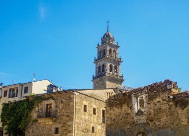 İspanya 'nın Ponferrada kentindeki Basilica de la Encina kulesi, açık mavi gökyüzünün altındaki tarihi taş binaların üzerinde yükseliyor. Yakınlarda yürüyen insanlar eski ve yeni mimarinin bir karışımını sergiliyor..