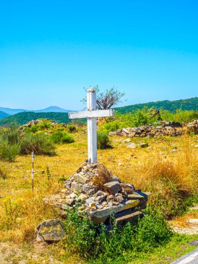 Camino de Santiago boyunca uzanan taşların üzerinde yıpranmış beyaz bir haç duruyor. Yuvarlanan tepeler ve berrak mavi gökyüzü, anıları ve ruhani yansımaları simgeliyor..