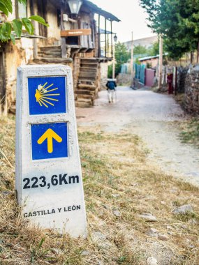 Camino de Santiago 'da kırsal bir yolda yürüyen bir seyyah ikonik sarı karabatak kabuğu ve sarı ok sembolleriyle bir dönüm noktasından geçiyor. Bu da Santiago de Compostela' ya 223.6 km kaldığını gösteriyor..