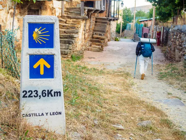 stock image A pilgrim walking along a rural road on the Camino de Santiago, passing by a milestone with the iconic Yellow Scallop Shell and Yellow Arrow symbols, indicating 223.6 km remaining to Santiago de Compostela in the region of Castilla y Leon.