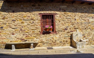 A rustic stone wall featuring a small barred window with pink flowers, a stone bench, and a water fountain on a sunny day in a traditional Spanish village. clipart