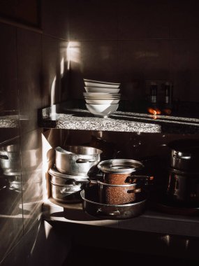 Loş bir mutfak köşesi dramatik güneş ışığının içeri aktığı istiflenmiş tabakları, çanakları, tencereleri ve tavaları aydınlattığı. Granit tezgahta çeşitli yiyecek ve hasır sepet bulunur..