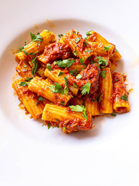 A close-up of Rigatoni pasta served with rich tomato sauce, garnished with fresh parsley. The dish is presented on a white plate, highlighting the vibrant colors and appetizing texture of the pasta.