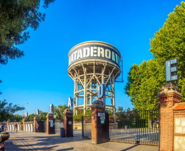 Madrid, Spain - September 3, 2024. The striking Matadero water tower stands tall against a vivid blue sky, framed by lush green trees. An entrance gate with brick pillars and iron fencing leads to this landmark of Madrid's cultural center. clipart