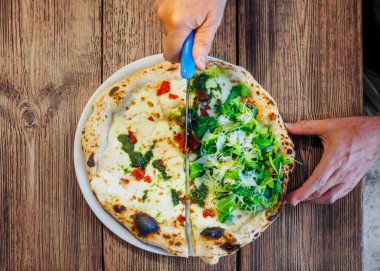 Bir yarısı erimiş peynir ve pesto soslu, diğer yarısı parmesan talaşlı taze roka yapraklı, sade ahşap bir masada servis edilen zanaatkar Napoli usulü pizza..