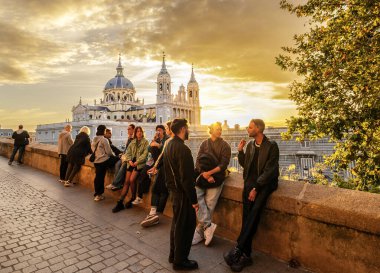 Madrid, İspanya. 1 Kasım 2024. Larra Garden 'dan gün batımında Almudena Katedrali' nin manzarası. Bir grup genç, ön planda Arap taş duvarının kalıntıları arasında toplandı..