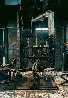 Endüstriyel atölyenin yakınında bir alet tahtası, anahtarlar, pense, zincirler, mekanik parçalar ve havalandırma borusu var. Yıpranmış zemin ve loş ışık, engebeli ve işlevsel atmosferini vurguluyor..