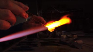Bir atölyedeki gaz lambasının üzerinde duran cam boncuğu şekillendiren ellerin yakın plan çekimi. Cam işi mücevherler.