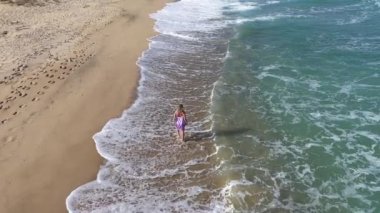 Yaz elbisesi giymiş, kumlu tropikal okyanus sahillerinde deniz dalgaları ve yaz tatili ile yürüyen genç bir kadının havada aktif takibi.