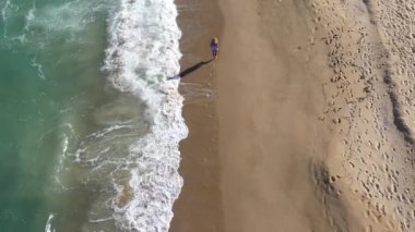 Kumsalda yürüyen muhteşem sarışın bir kadının hava manzarası. Deniz dalgaları kumlu kıyı şeridini yıkıyor