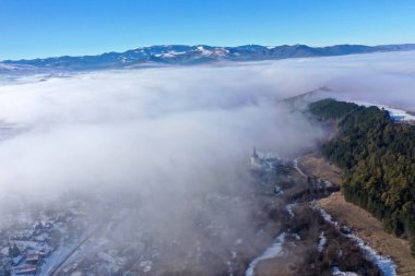 Aerial view of foggy misty mountain village and church tower at winter by drone clipart