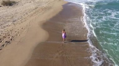Kumsalda ağır çekimde yürüyen sarışın model kadın. Deniz dalgaları kumlu sahili İHA 'larla yıkıyor.