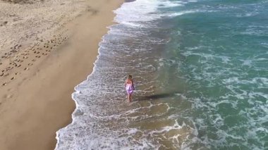 Kumsalda ağır çekimde yürüyen sarışın model kadın. Deniz dalgaları kumlu sahili İHA 'larla yıkıyor.