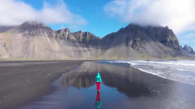  Atlantik Okyanusu kıyısında koşan bir kadının havadan yavaş çekim görüntüsü. Siyah kumsalları yıkayan deniz dalgaları. Hofn, Vestrahorn Dağları, İzlanda