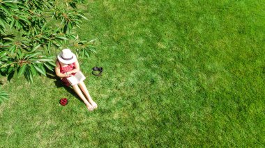 Parkta kitap okuyan genç bir kadın, dışarıda otların üzerinde kitap ve kulaklıklarla rahatlayan bir öğrenci kız, yukarıdan hava aracı manzaralı.