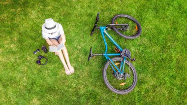 Bisikletin yanındaki parkta kitap okuyan genç bir kadın. Öğrenci kız dışarıda çimenlerde oturuyor bisiklet, kitap ve kulaklıkla oturuyor, yukarıdan insansız hava aracı görüşü