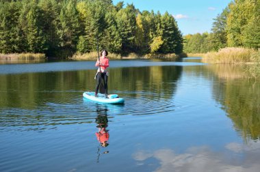 Active woman paddling SUP board on beautiful lake, autumn forest landscape and nature on background, stand up paddling water adventure outdoors, sport and healthy lifestyle concept