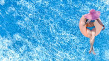 Yüzme havuzunda şapkalı güzel bir kadın yukarıdan drone manzaralı, bikinili genç bir kız şişme yüzüklü çöreklerle rahatlıyor ve tatilde tropikal tatillerde suda eğleniyor.