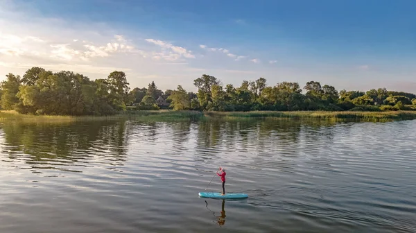 Woman paddling on SUP board on beautiful lake aerial drone view with reflections from above. Standing up paddle boarding adventure in early morning sunrise. Germany lake district Mecklenburg.