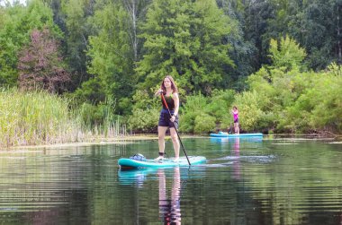 Active family on SUP, mother and daughter paddling on standing up paddleboards in river water, summer family sport on supboards clipart