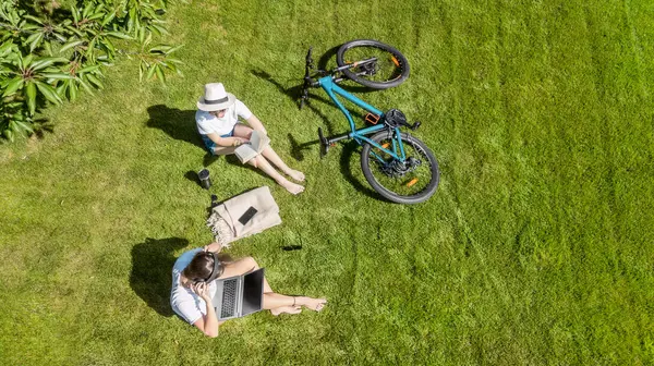 Parkta bisikletli genç kızlar, dizüstü bilgisayar ve kulaklık kullanıyorlar, ders çalışan ve dışarıda dinlenen iki kız öğrenci bisiklet yakınındaki çimlerde oturuyor, yukarıdan drone görüntüsü var.