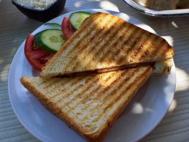 Lezzetli bir ızgara jambonlu ve peynirli tost sandviçi yanında taze salatalık ve domates dilimleri ile servis edilir. Çıtır çıtır, altın sarısı ekmek, erimiş peynir ve jambonla mükemmel bir şekilde kızartıldı. Klasik ve basit bir mola