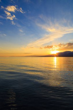 Deniz ve dağların üzerinde güzel bulutlu gün batımı. Turuncu güneşin ışınlarında parlak bir gökyüzü. Doğal
