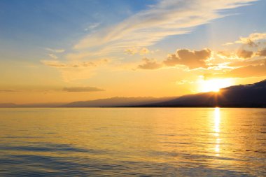 Deniz ve dağların üzerinde güzel bulutlu gün batımı. Turuncu güneşin ışınlarında parlak bir gökyüzü. Doğal