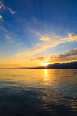 Deniz ve dağların üzerinde güzel bulutlu gün batımı. Turuncu güneşin ışınlarında parlak bir gökyüzü. Doğal