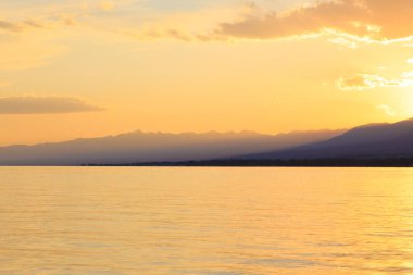 Deniz ve dağların üzerinde güzel bulutlu gün batımı. Turuncu güneşin ışınlarında parlak bir gökyüzü. Doğal