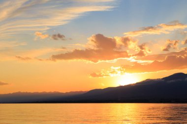 Deniz ve dağların üzerinde güzel bulutlu gün batımı. Turuncu güneşin ışınlarında parlak bir gökyüzü. Doğal
