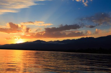Deniz ve dağların üzerinde güzel bulutlu gün batımı. Turuncu güneşin ışınlarında parlak bir gökyüzü. Doğal