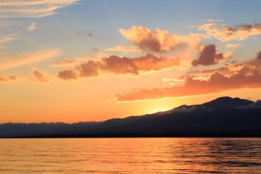 Deniz ve dağların üzerinde güzel bulutlu gün batımı. Turuncu güneşin ışınlarında parlak bir gökyüzü. Doğal